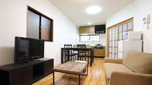 Uma sala de estar com televisão, mesa de centro de madeira, sofá e área de jantar com cadeiras, e uma cozinha ao fundo.