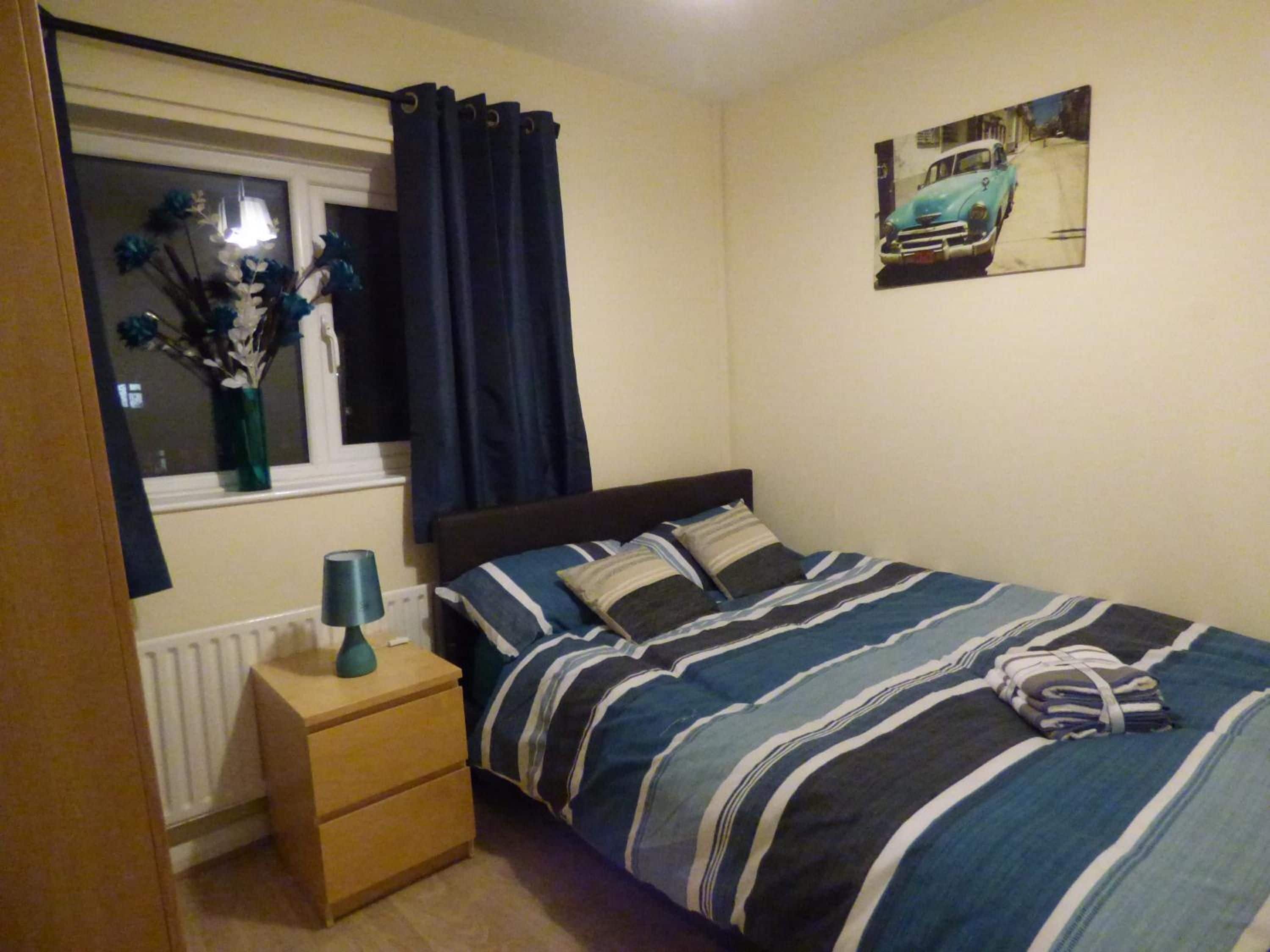 Une chambre à coucher comprenant un lit, une table de chevet, une fenêtre avec des rideaux, un radiateur et un tableau accroché au mur.
