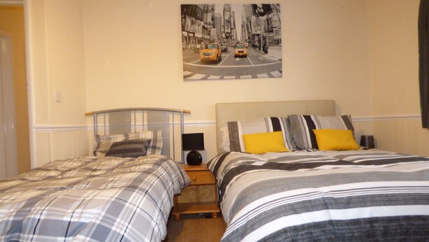 A hotel room with two beds, a nightstand, a lamp, and a cityscape painting on the wall. - Beckley House (Nottingham)