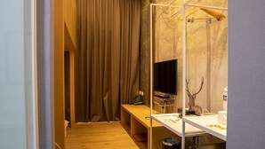 A compact hotel room with a wooden bench, a desk with a computer, a mirror, and a deer head decoration.