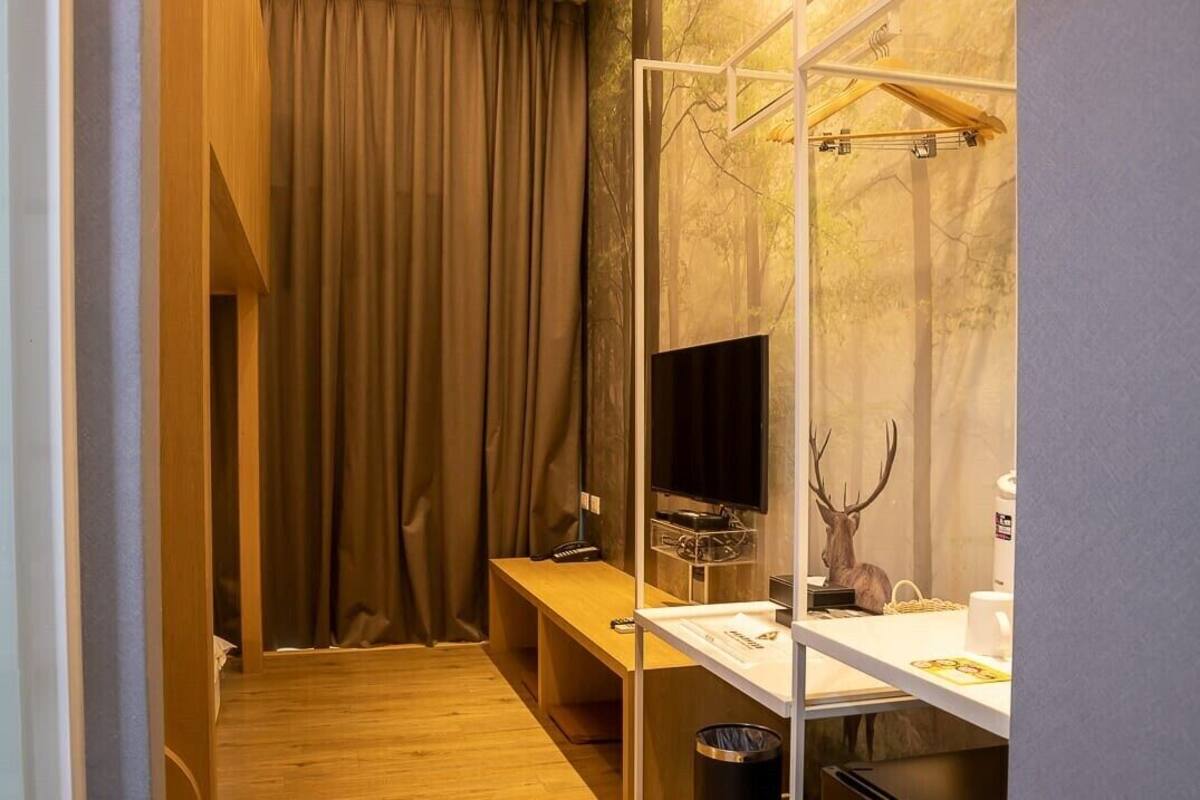 A compact hotel room with a wooden bench, a desk with a computer, a mirror, and a deer head decoration.