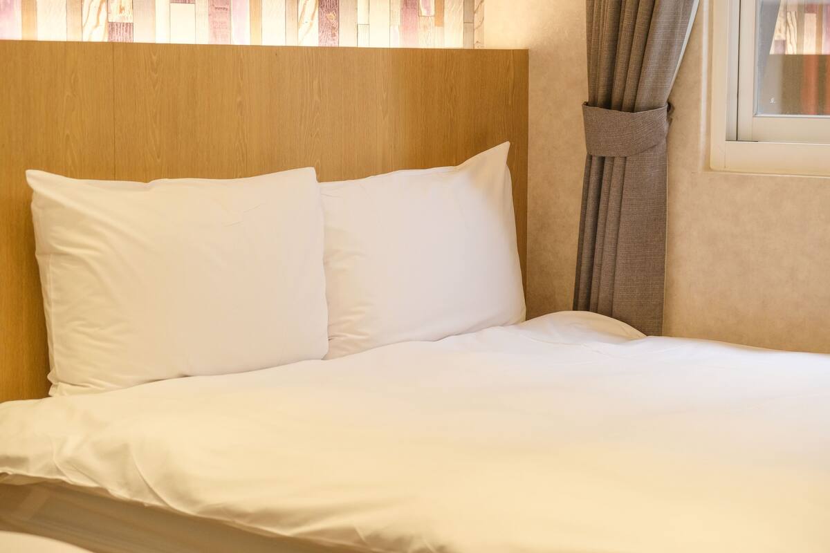 A neatly made bed with white linens and pillows, a wooden headboard, and a window with curtains.