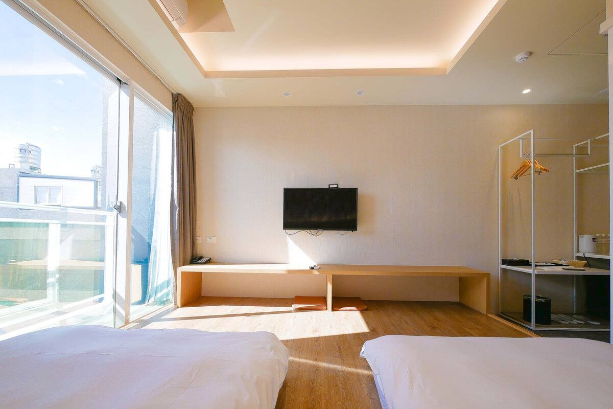 A hotel room with two beds, a TV mounted on the wall, a desk, and a large window offering a view of the city.