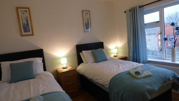 Een hotelkamer met twee bedden, elk voorzien van nachtkastje en lamp. Er hangen prestatiekaders aan de muren en er is een raam met gordijnen.
