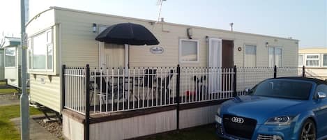 A mobile home with a fenced patio area, a parked blue car, and a small table with chairs.
