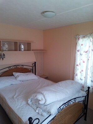 Une chambre avec un lit simple, une table de chevet, une étagère avec des objets décoratifs et une fenêtre ornée de rideaux à motifs floraux.