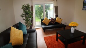 A living room with a black sofa, yellow and teal cushions, a small table with a vase of flowers, and a large window with curtains. - Depedale House (Derby)