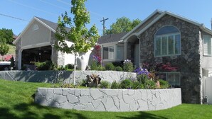 Una casa a due piani con facciata in pietra, un garage e un giardino piantumato con diverse piante e fiori.