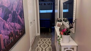 Un corridoio con un tappeto decorato, un quadro di grandi dimensioni, un consolle bianco con uno specchio e una porta che dà su una camera da letto.