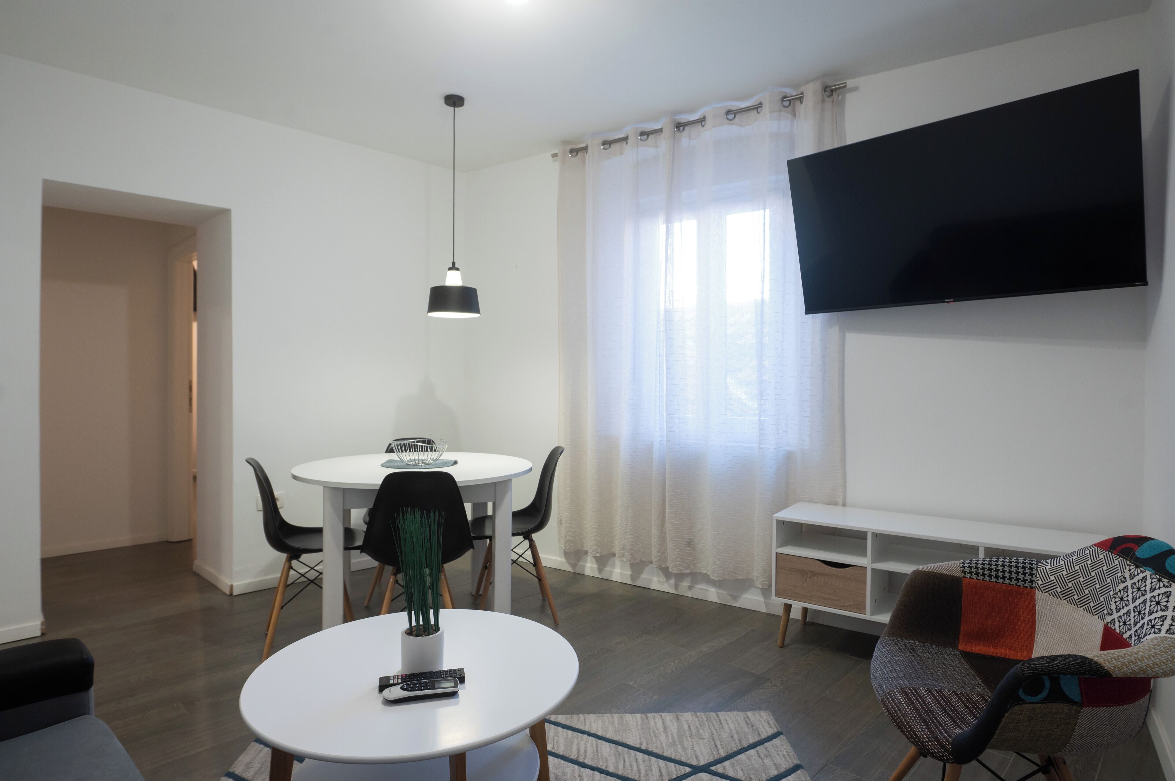 Ein modernes Wohnzimmer mit einem runden Esstisch, zwei Stühlen, einer Couch, einem an der Wand montierten Fernseher und einem großen Fenster mit Vorhängen.