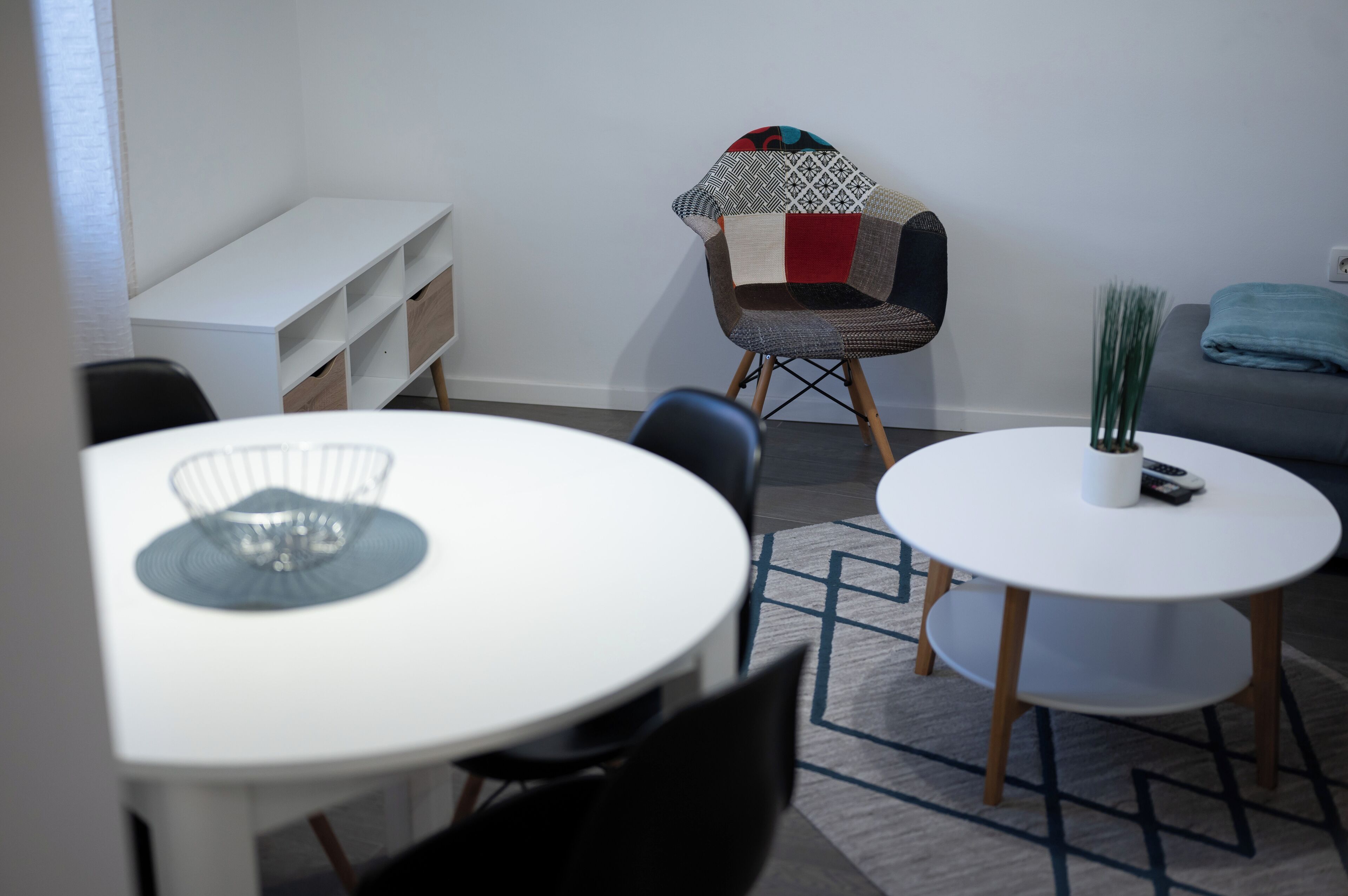 Una habitación moderna con un escritorio blanco, dos mesas redondas blancas, un sillón estampado y un sofá gris.