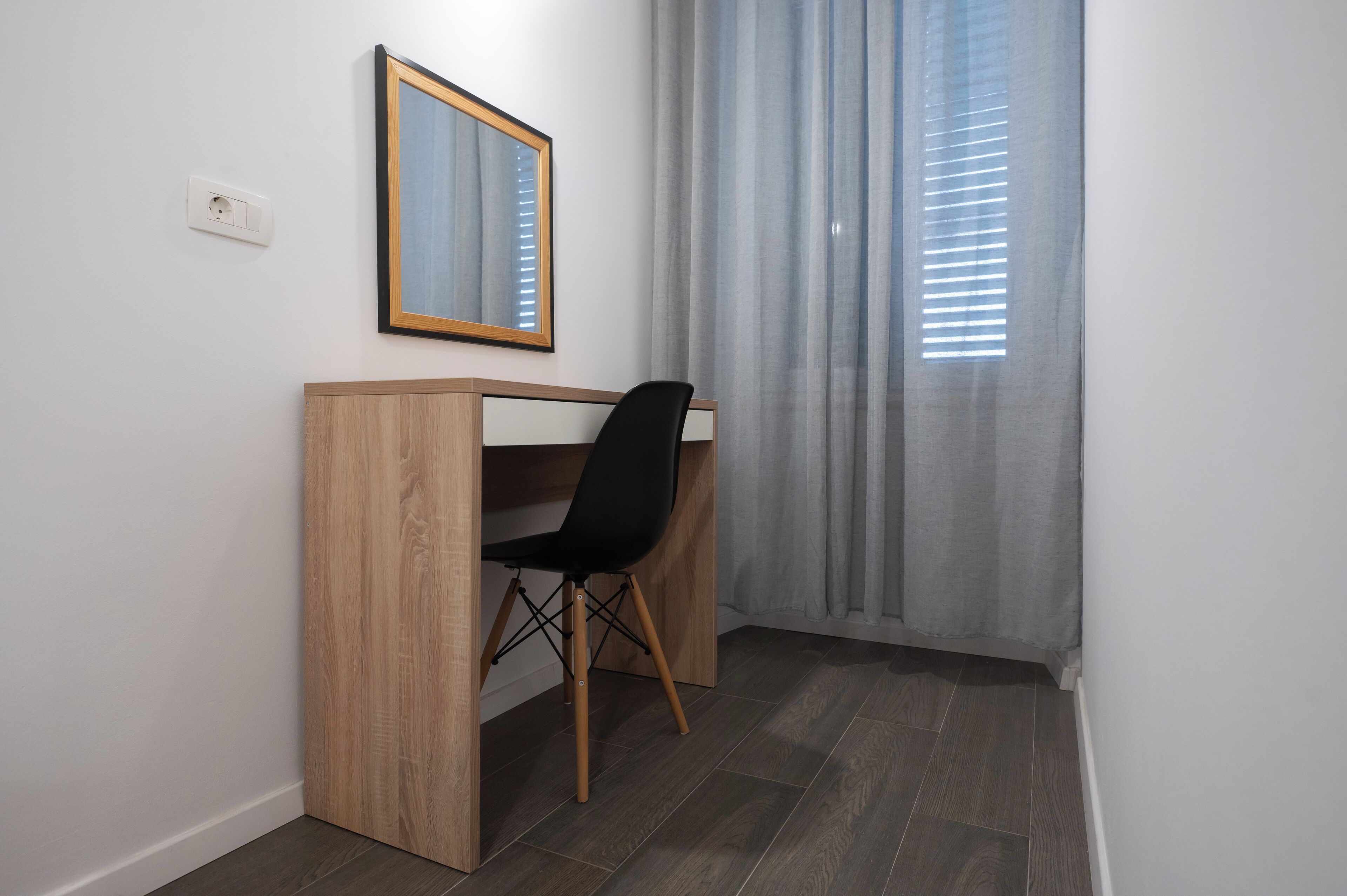 A small office space with a wooden desk, a black chair, a mirror, and a window with curtains.