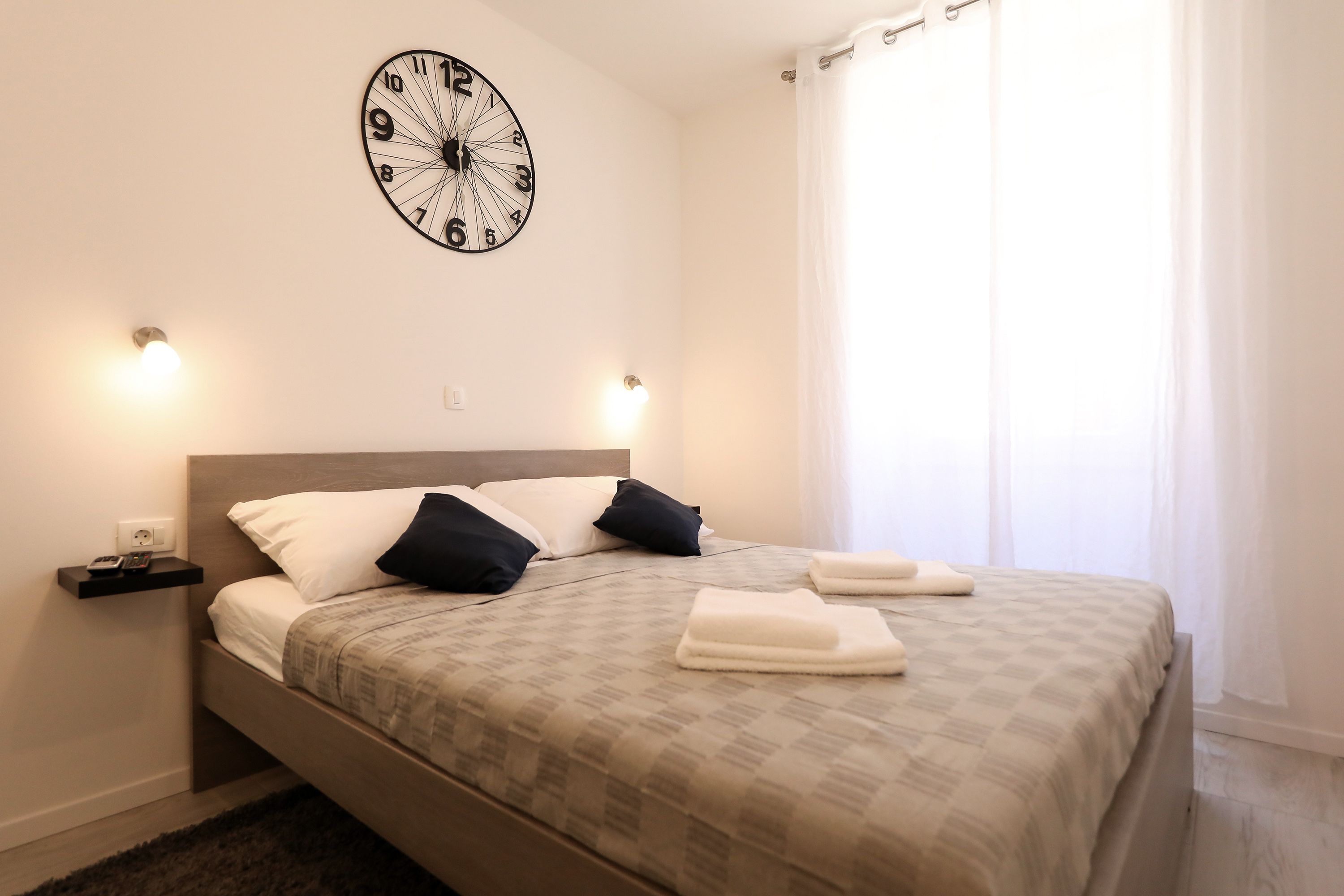 A neatly made bed with a patterned bedspread, two pillows, and folded towels on a headboard. A wall clock is mounted above the bed, and there are two wall-mounted lights on either side.