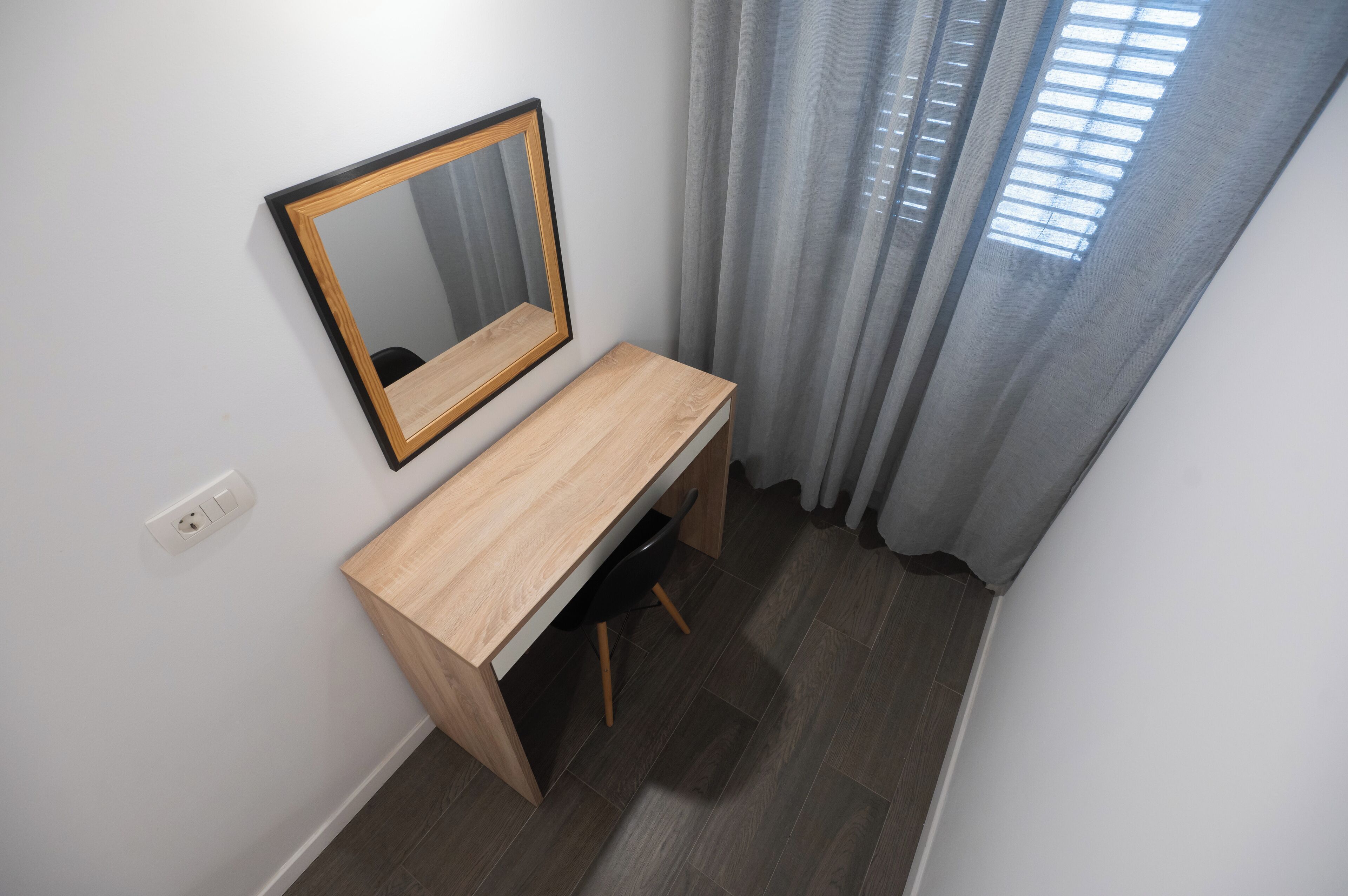 Una habitación pequeña y ordenada con un escritorio de madera, un espejo y una ventana con cortinas.