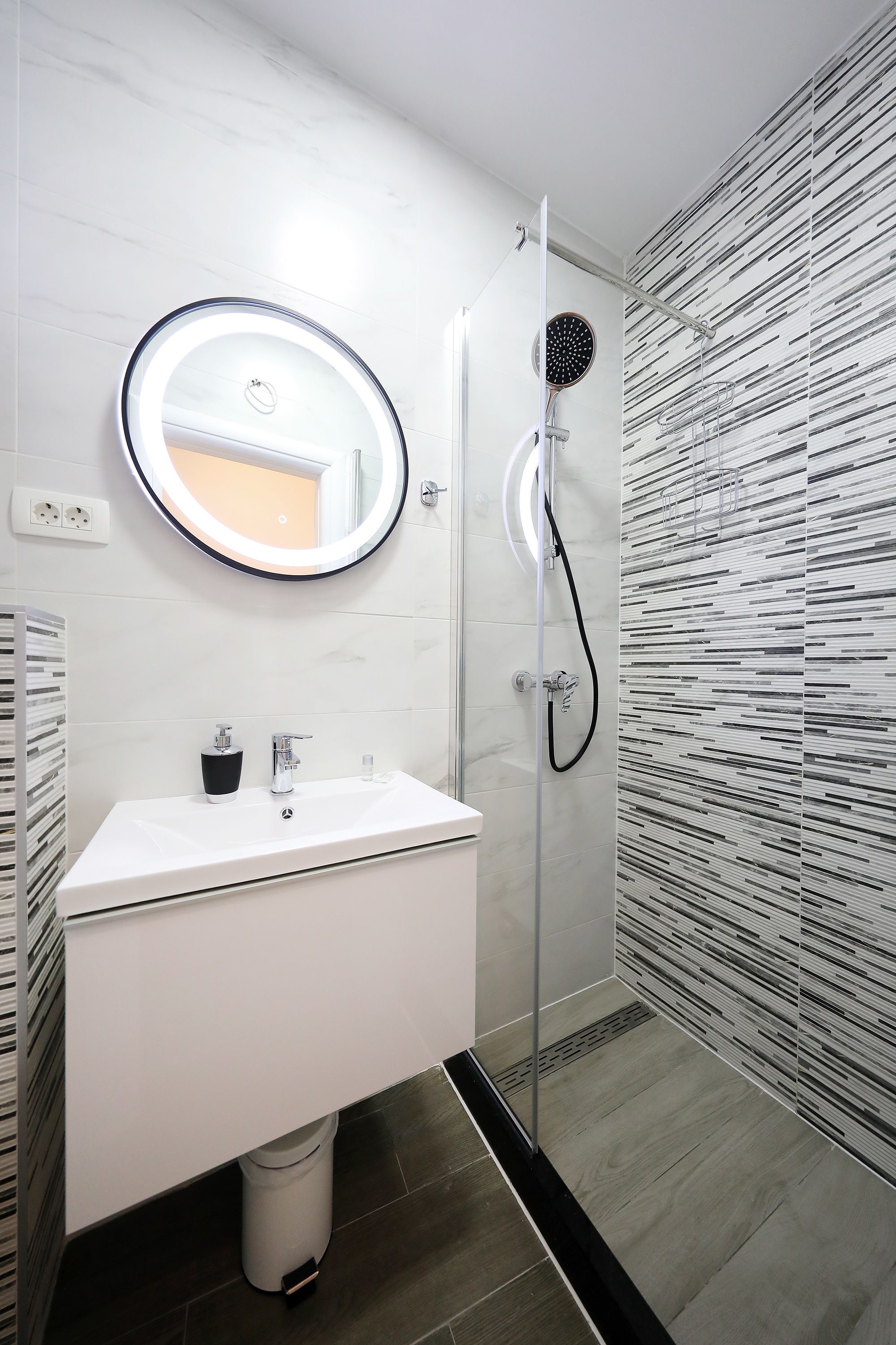 A modern bathroom with a white vanity, a round mirror, a handheld showerhead, and a patterned wall.