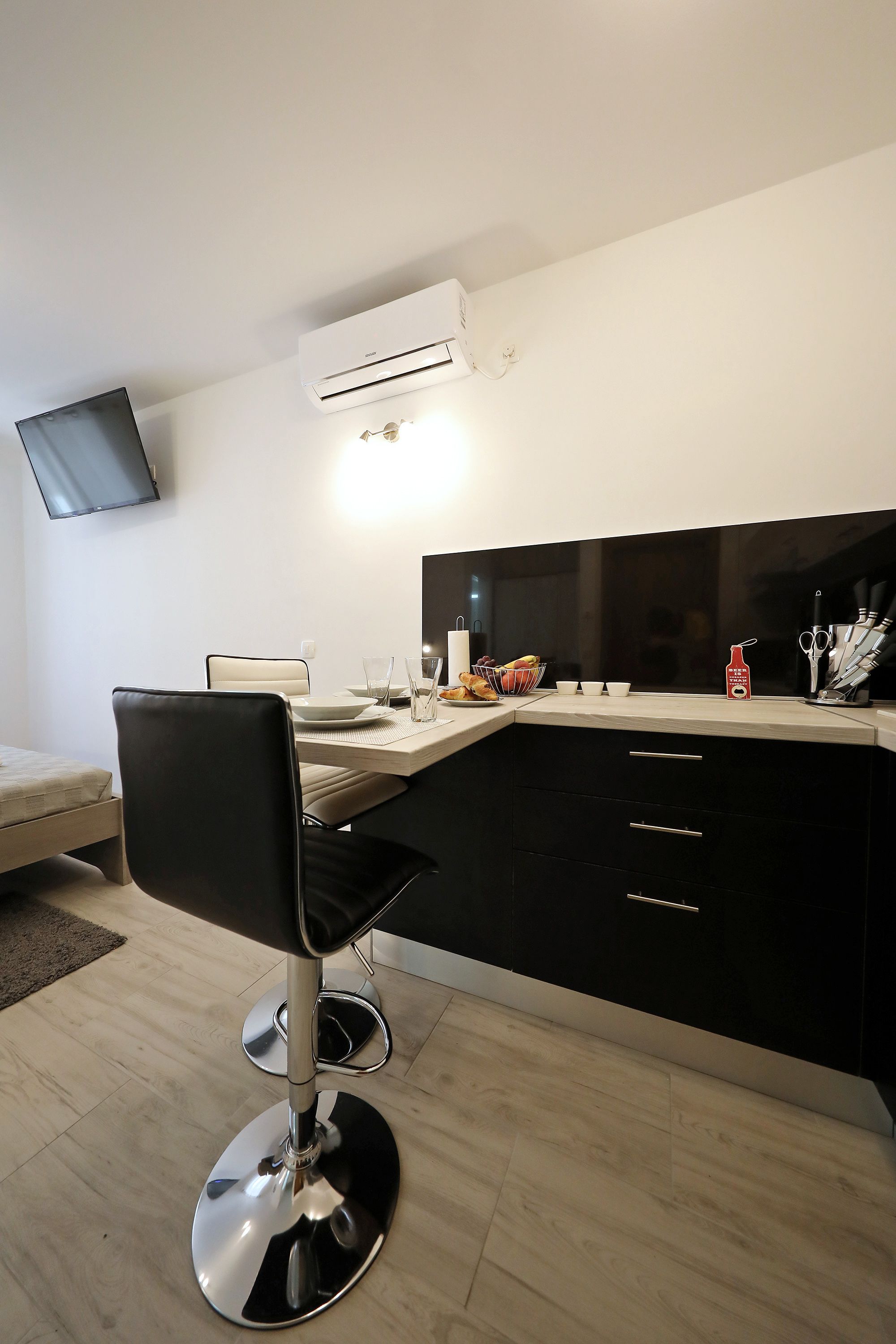 A modern kitchen with a dining area, including a black chair, a TV mounted on the wall, and an air conditioning unit.