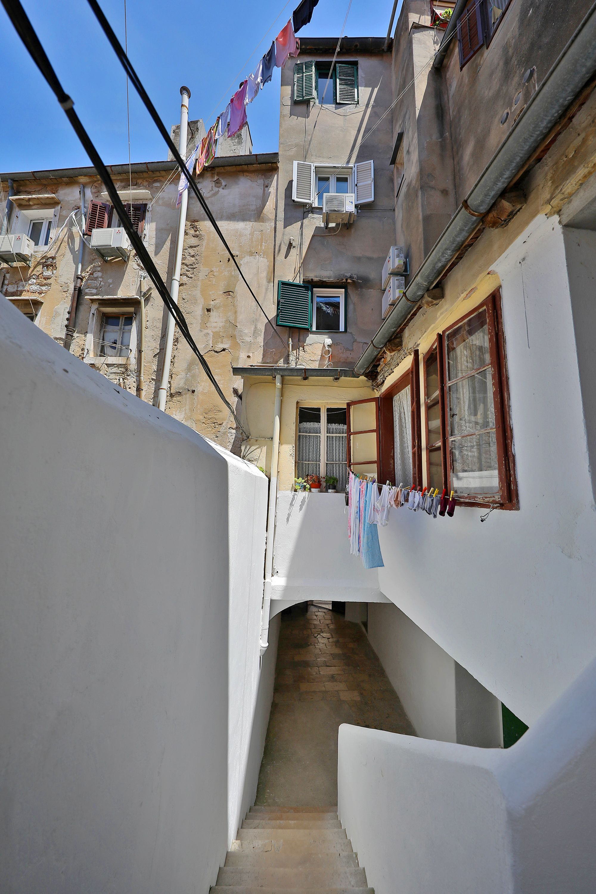 A narrow alleyway with a staircase leading to a building with multiple windows and air conditioning units.