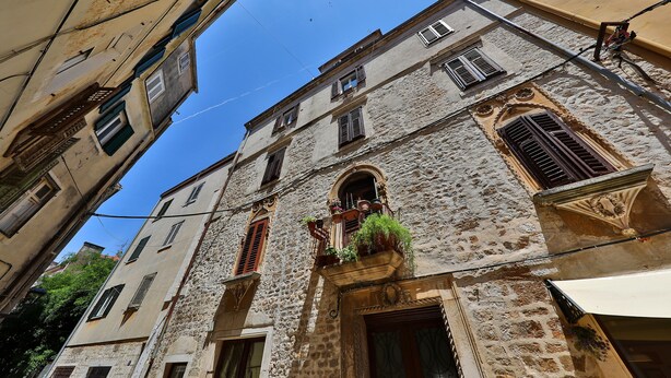 Una calle estrecha con edificios históricos de piedra, uno con un balcón adornado con plantas.