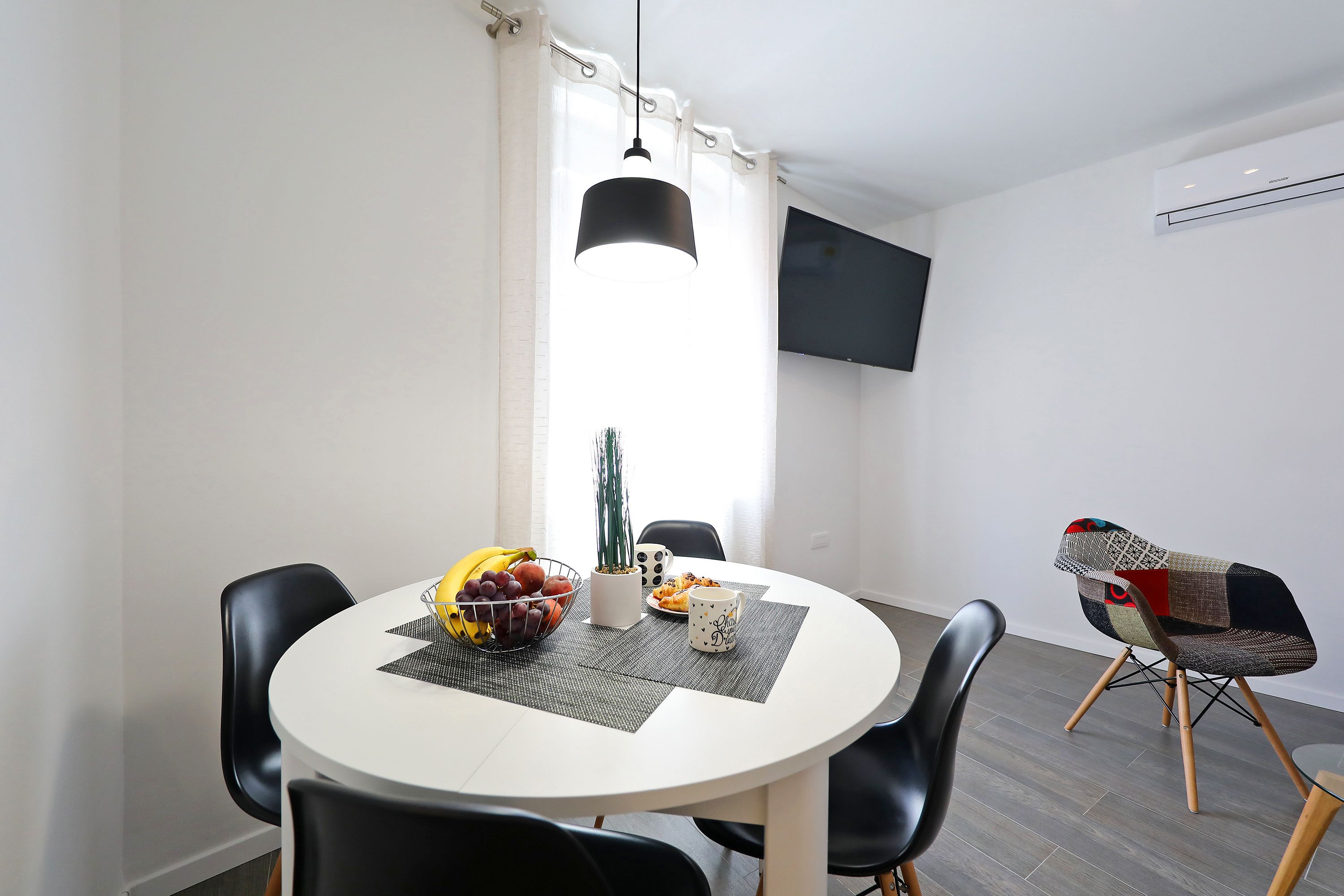 A modern dining area with a round table, black chairs, a fruit bowl, a cactus, and a TV mounted on the wall.