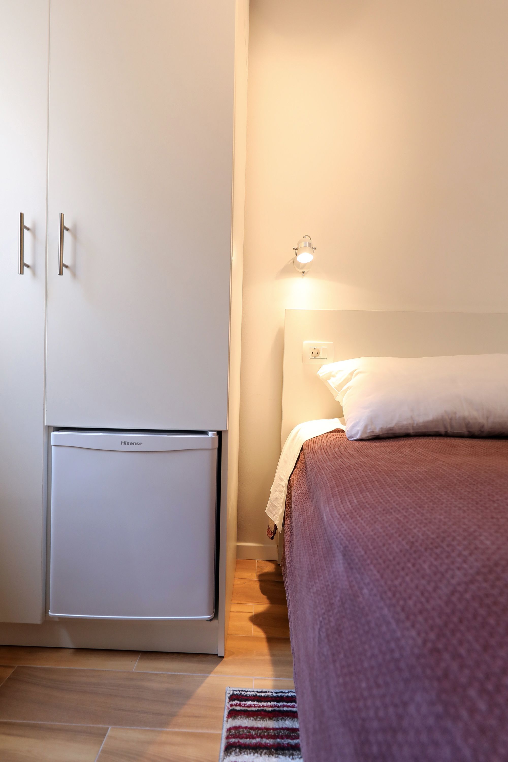 A bedroom with a bed, a bedside table, a nightstand, a wardrobe, and a small refrigerator.