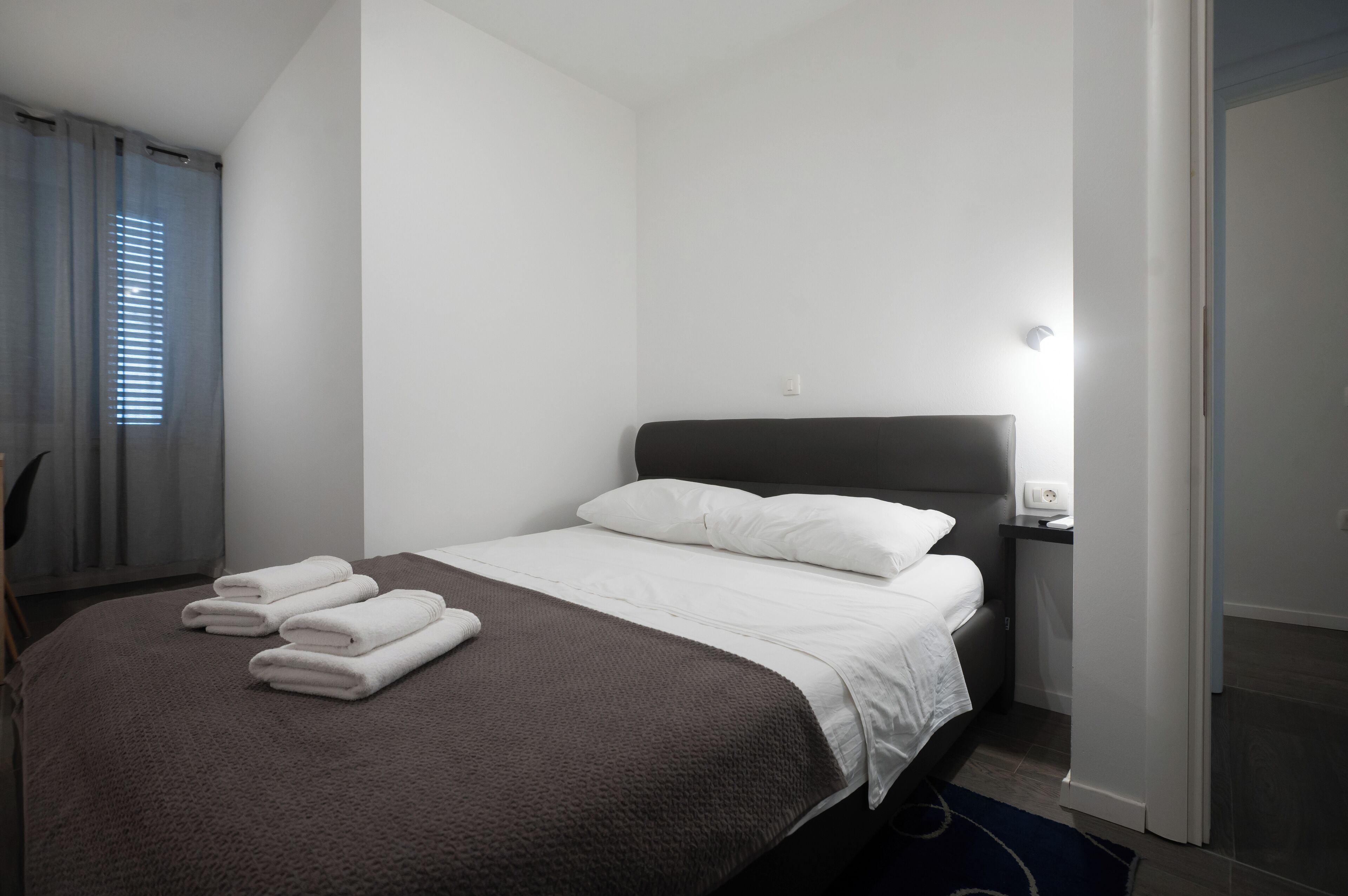 A neatly made bed with white linens and a brown blanket, a bedside table with a lamp, and a window with blinds.