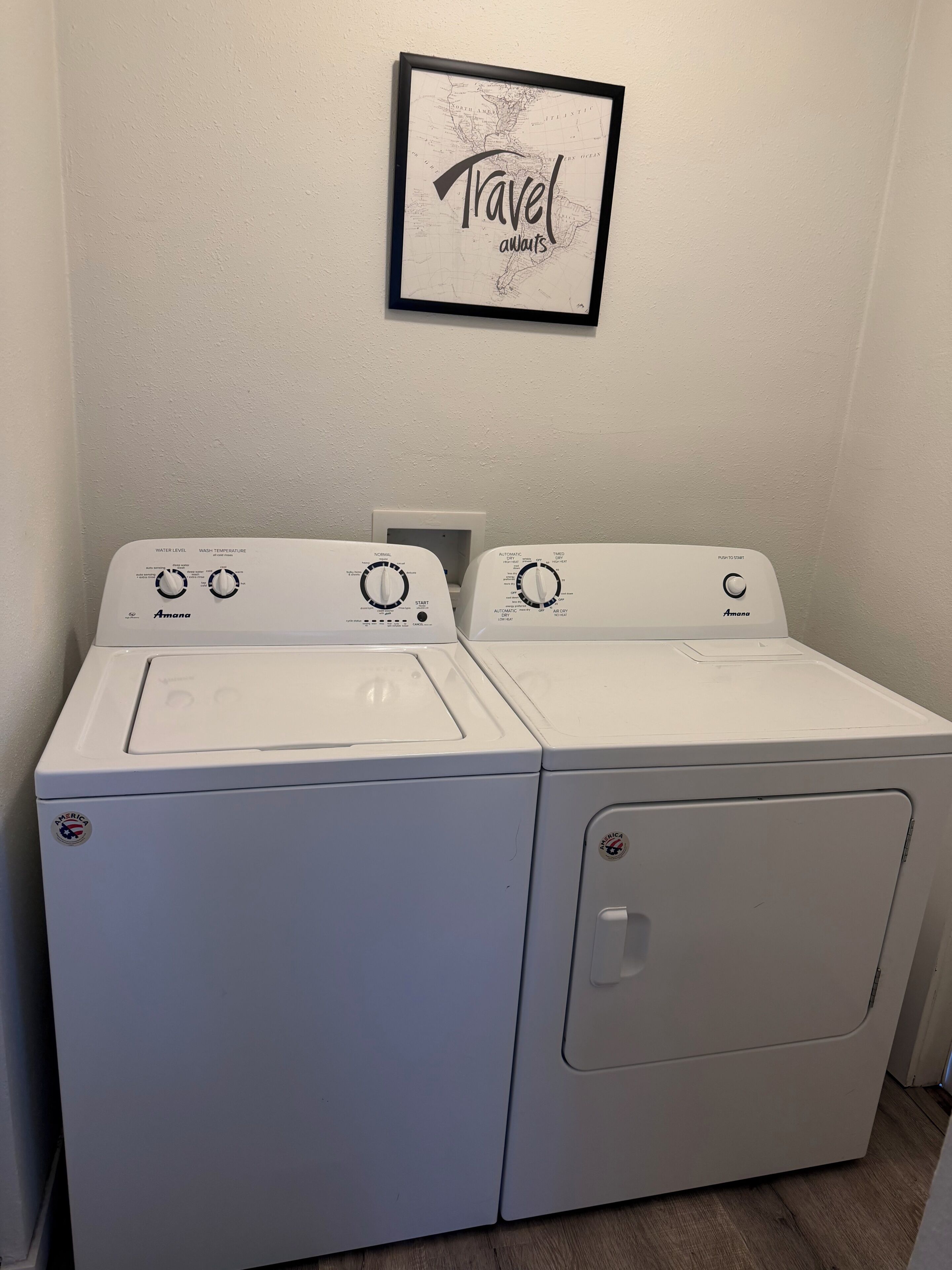 Een wasruimte met twee witte Amana wasmachines en drogers, een inlijsting kaart met de tekst 'Travel always' aan de wand, en een witte muur.