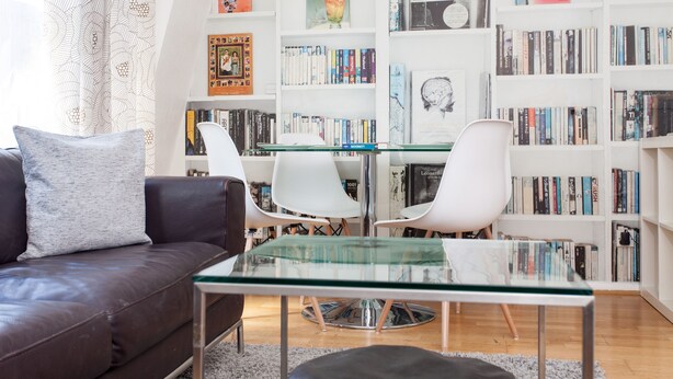 Ein modernes Wohnzimmer mit einem Glastisch, weißen Stühlen, einem braunen Ledersofa und einem Bücherregal voller Bücher.