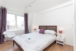 A neatly made bed with pillows, a bedside table with a lamp, a rocking chair, and a window with curtains.