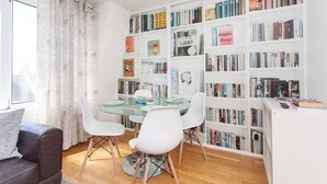Ein modernes Wohnzimmer mit einem Glastisch, weißen Stühlen, einem Bücherregal voller Bücher und einem großen Fenster mit Vorhängen.