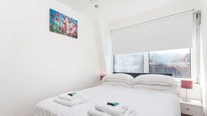 Ein ordentlich bezogenes Bett mit weißen Leinen und Handtüchern, ein Nachttisch mit Lampe und ein Fenster mit Blick auf Gebäude.