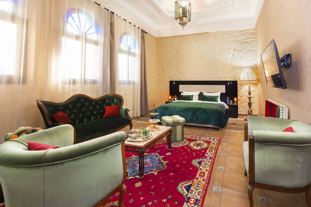 A hotel room with a large bed, two armchairs, a small table with a tray of items, a patterned rug, and a large window with curtains.
