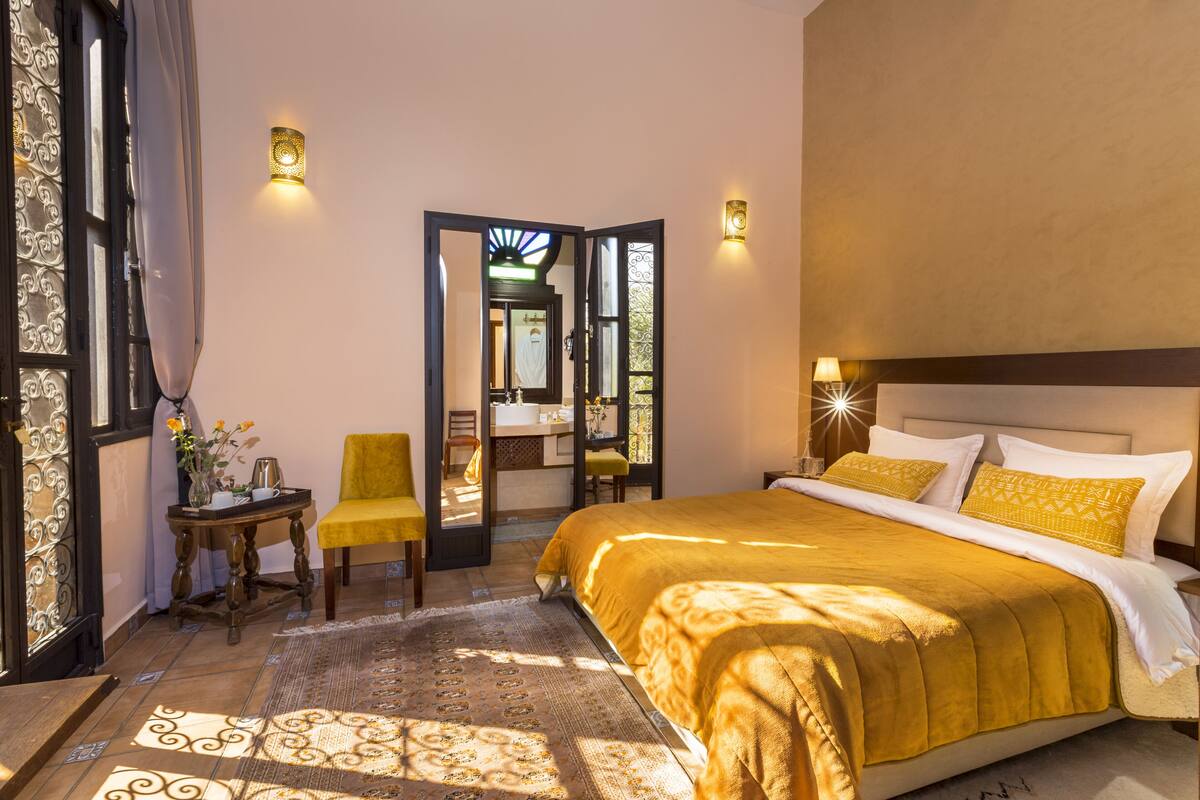 A hotel room with a large bed, a small table with a vase of flowers, a chair, and a view through an open door.