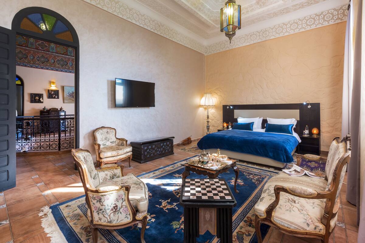 A hotel room with a large bed, two armchairs, a small table, a television, and a decorative archway.