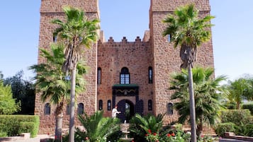 A stone castle with two towers, a central entrance, and a fountain surrounded by palm trees and flowers.
