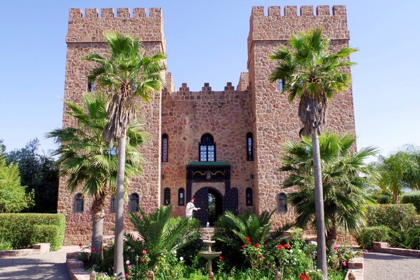 A stone castle with two towers, a central entrance, and a fountain surrounded by palm trees and flowers. - La Citadelle de Marrakech (Ghmate)