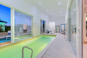 Une piscine intérieure moderne avec des murs en verre, un espace détente et une vue sur l’extérieur.