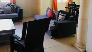 Una sala de estar con sofá, sillones, una mesa de comedor y una silla. Hay dos ventanas con cortinas, una lámpara de pared y un cuadro en la pared.