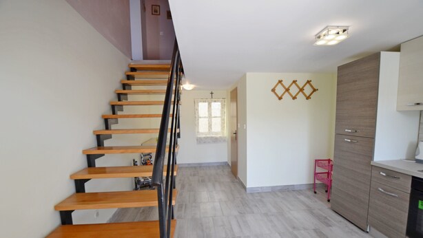 Una escalera de madera con barandillas negras que conduce a una habitación con zona de cocina y una pequeña silla roja.