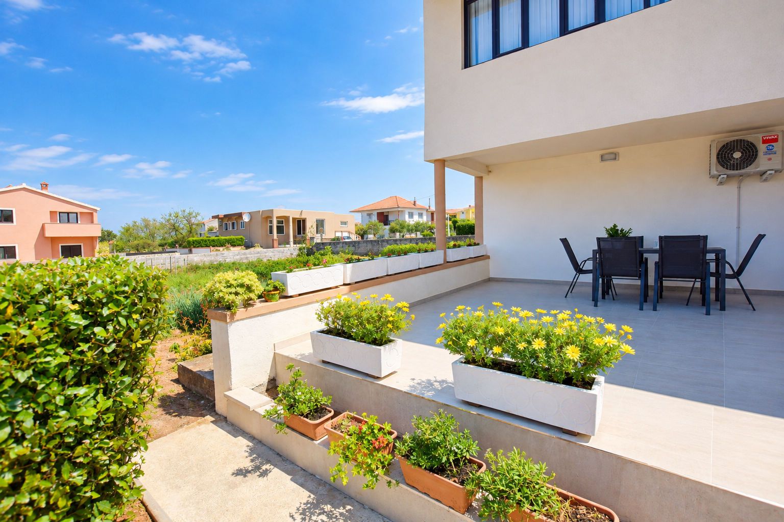 A modern outdoor patio with a dining area, planters with flowers, and a view of neighboring houses.