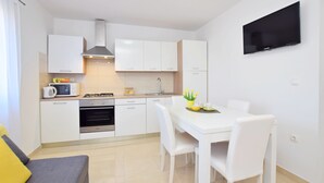 A modern kitchen with white cabinets, a microwave, stove, and a dining area with a white table and chairs. - Apartment Maja (Vrsi)