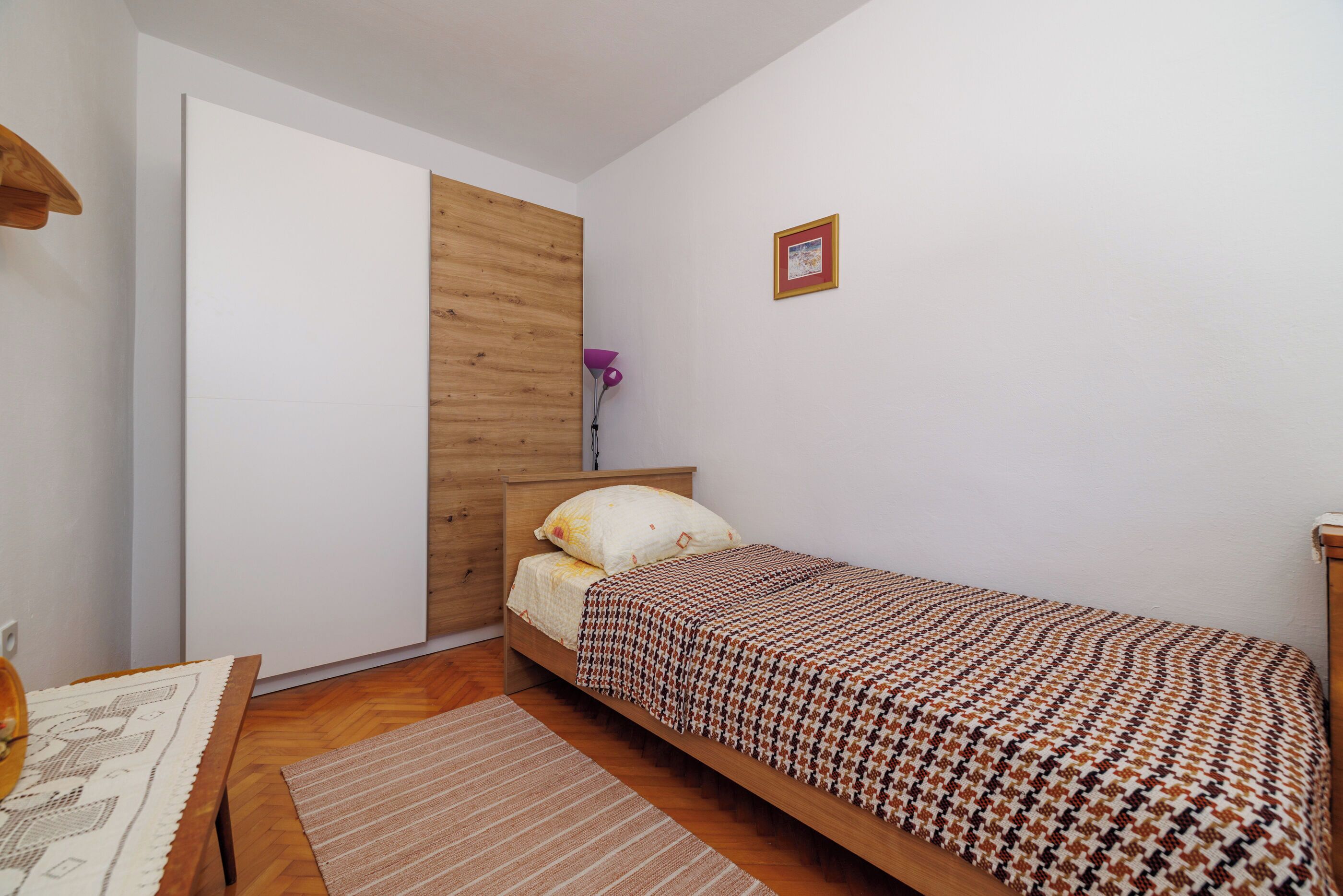 A small, single-bed room with a wooden headboard, a bedside table, a lamp, and a small picture on the wall.