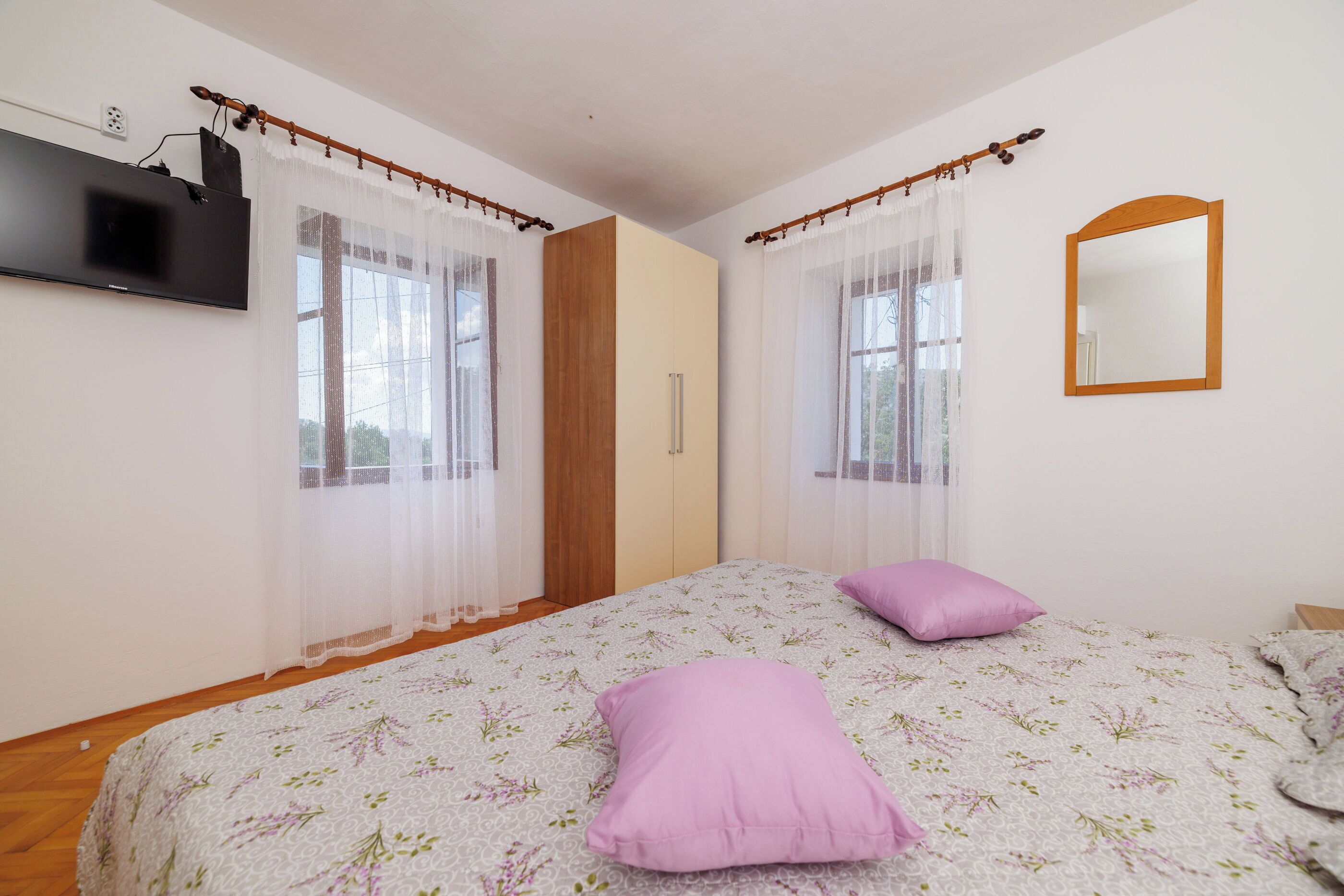 A bedroom with a bed, two pillows, a TV, a mirror, and a window with curtains.