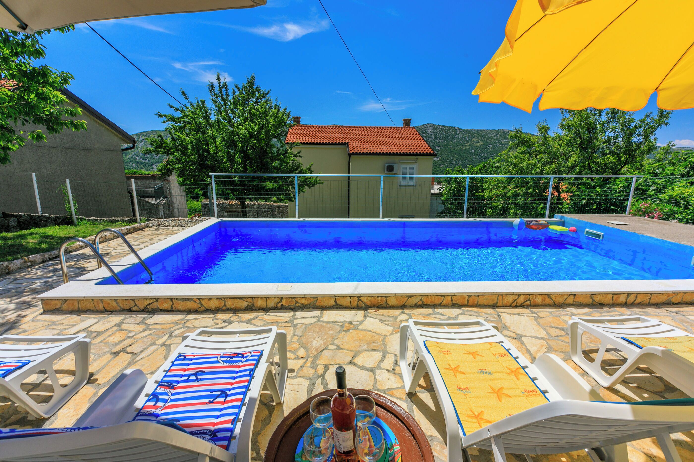 Piscina con poltrona prendisole e tavolino con bevande, circondata da piastrelle in pietra e vista sulle montagne.