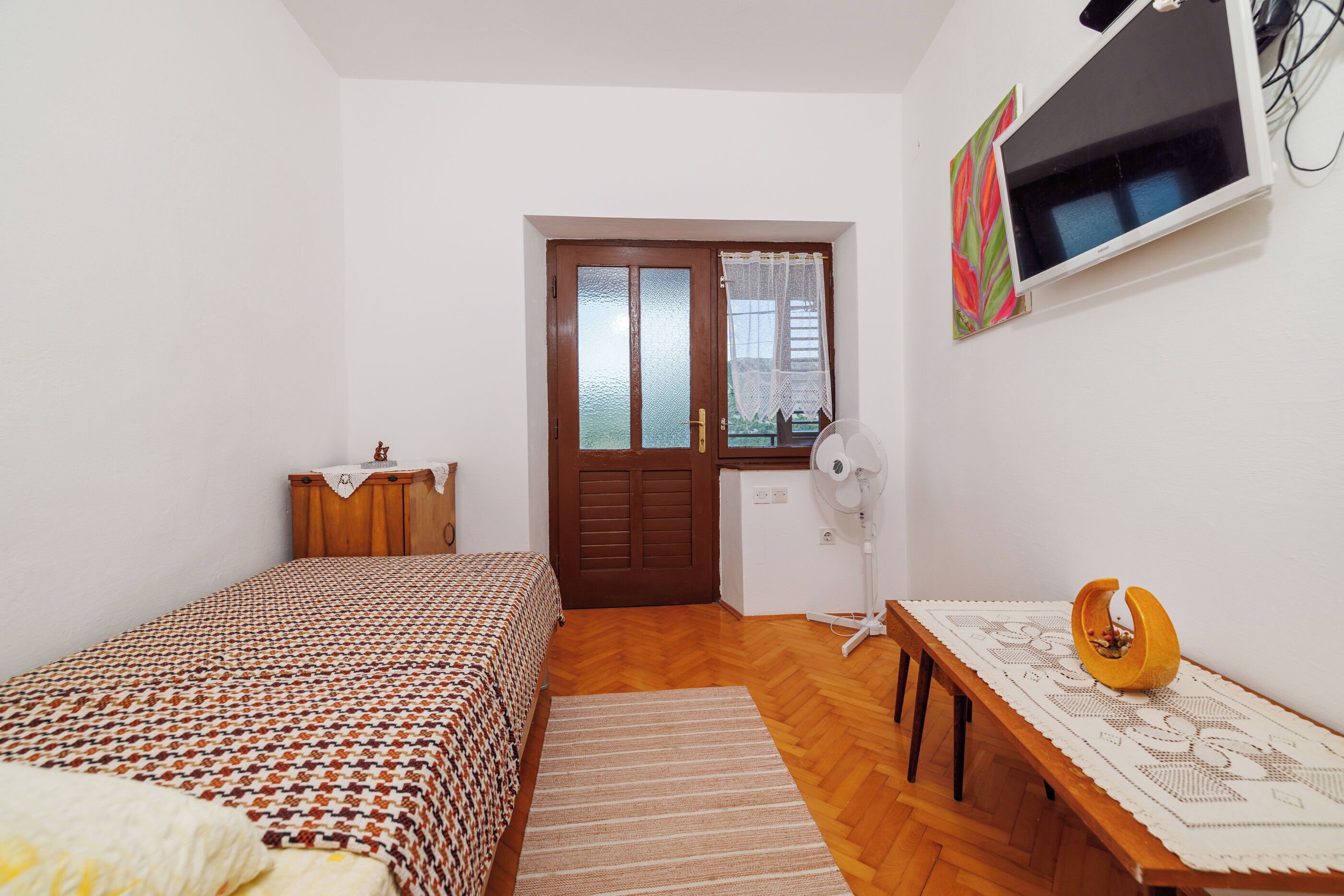 Un dormitorio con una cama, un aparador de madera, un escritorio con un ventilador, un televisor y una puerta con ventanal.