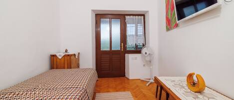 Un dormitorio con una cama, un aparador de madera, un escritorio con un ventilador, un televisor y una puerta con ventanal.