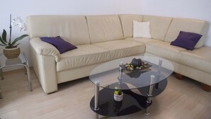 A beige sectional sofa with purple cushions, a glass coffee table with a vase and decorative items, and a potted orchid.