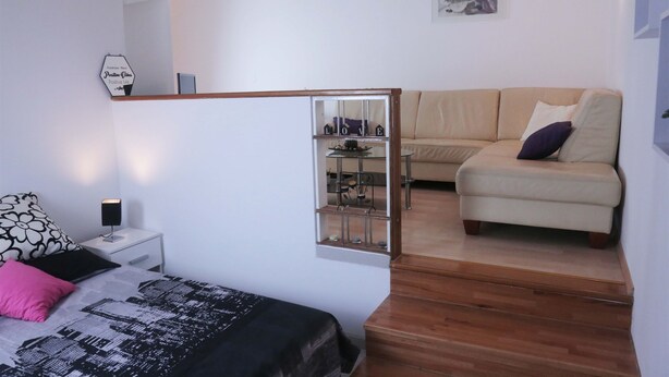 A bedroom with a bed, a nightstand, a dresser, a mirror, a wooden staircase, a sofa, and a wall-mounted shelf with decorative items.