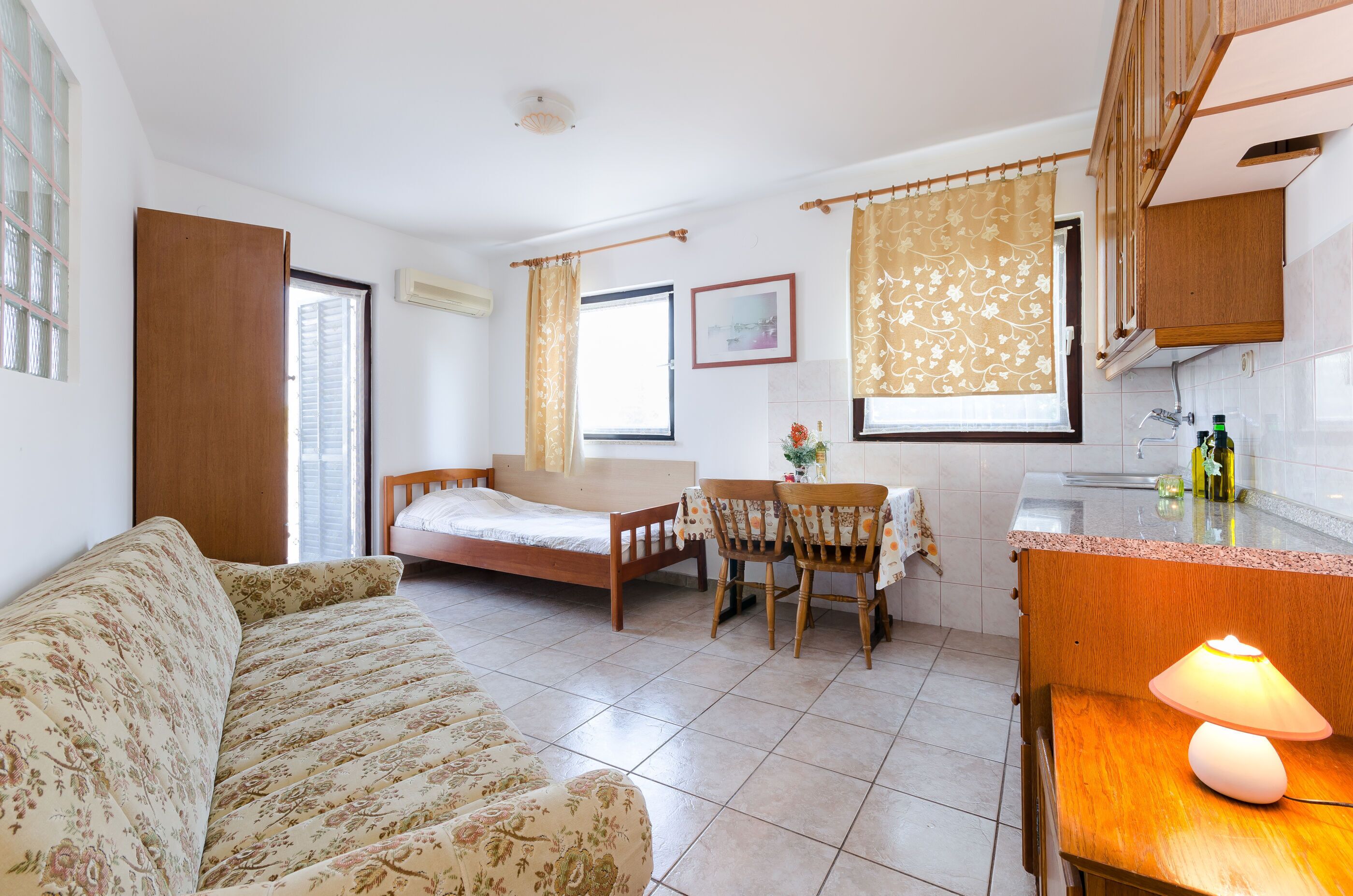 A compact room with a bed, a sofa, a dining table with chairs, a kitchen area, and a lamp.