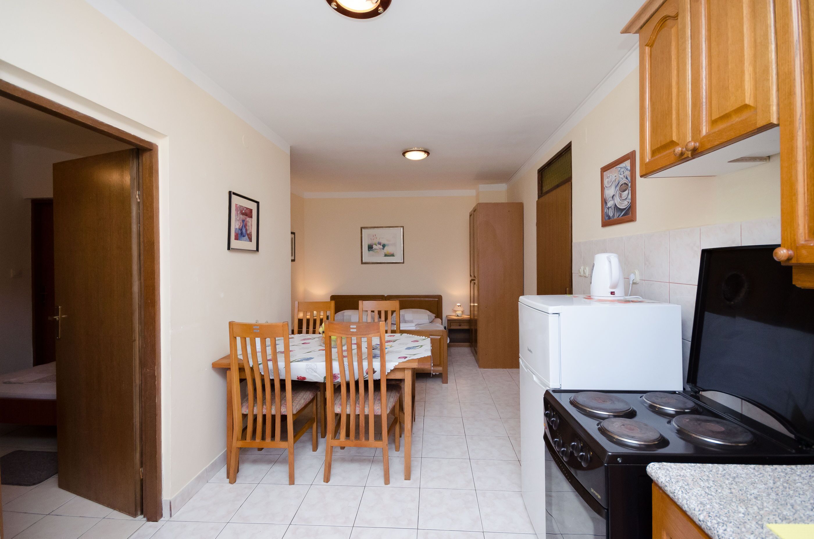 A compact kitchen with a stove, a refrigerator, wooden chairs, a dining table, and a bed in the background.