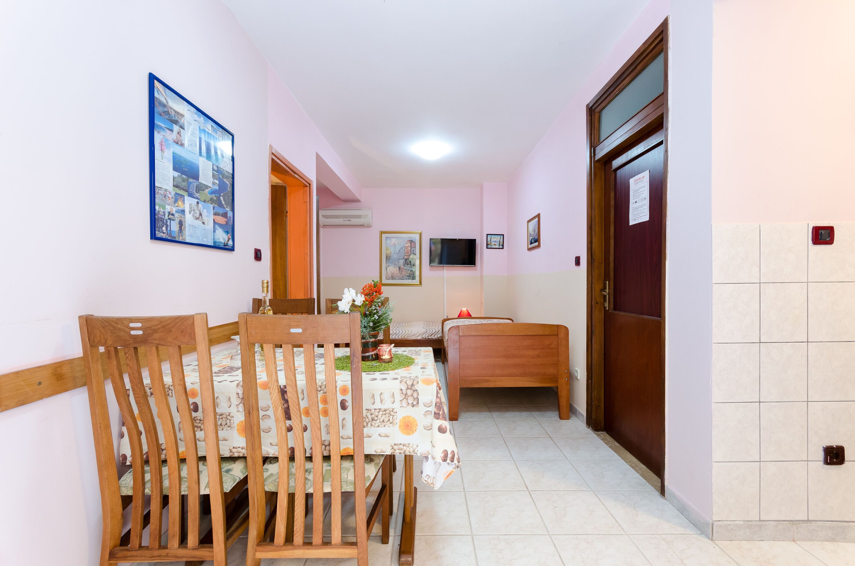 A dining area with a table set for two, wooden chairs, a small plant, and a wall-mounted TV.
